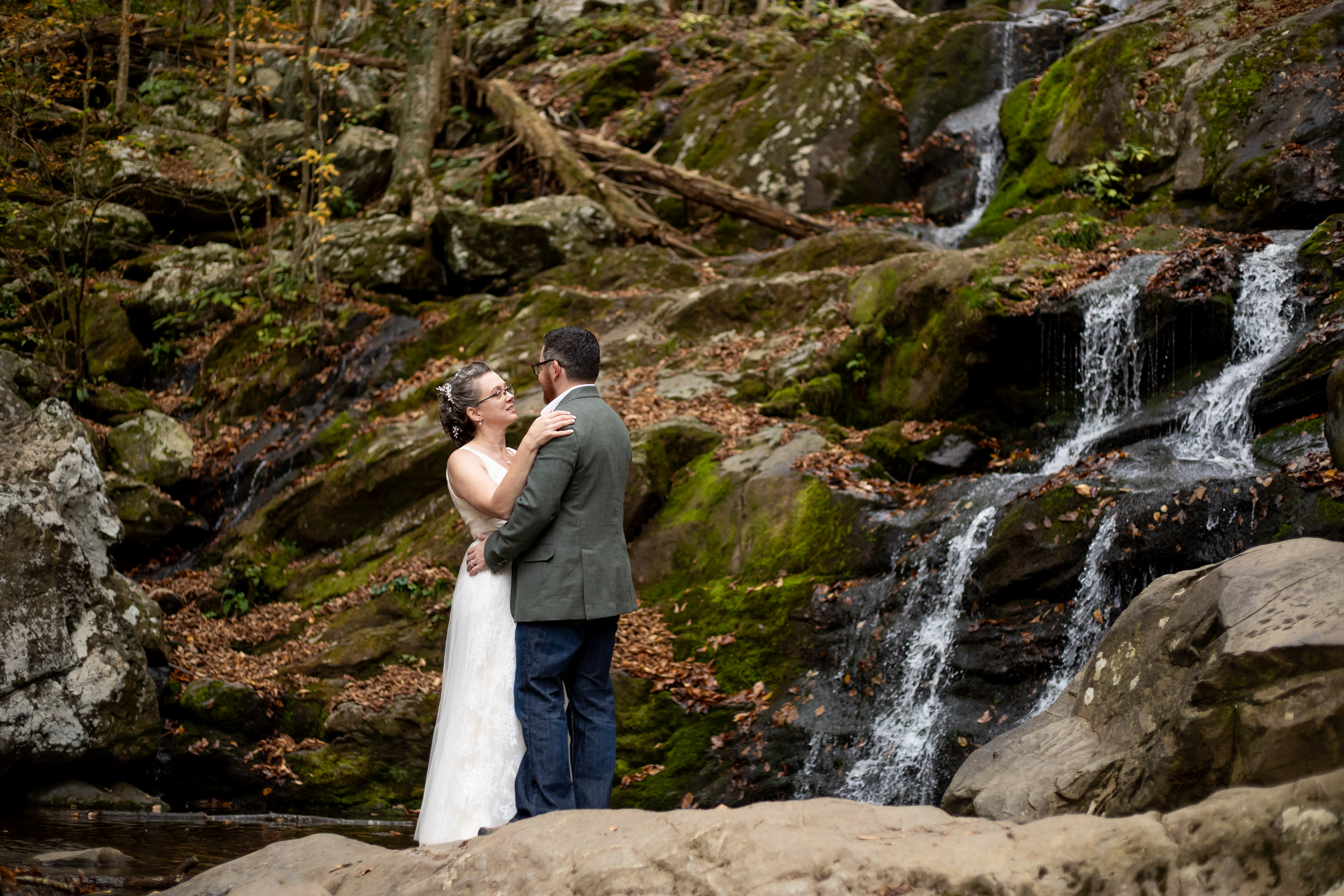 Couple embracing at the base of Dark Hollow Falls in Shenandoah National Park, surrounded by moss-covered rocks and cascading water