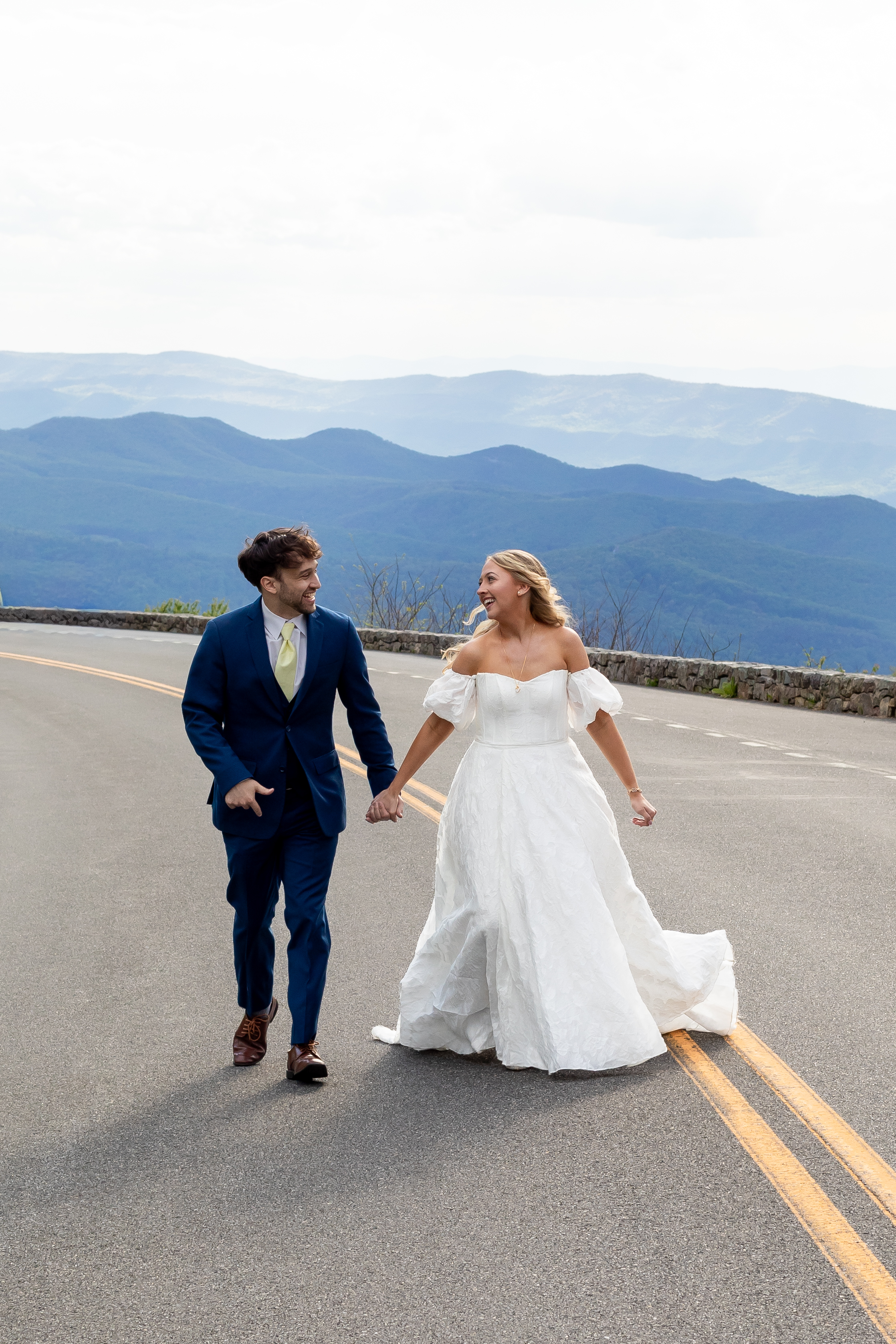 Couple laughing and walking hand in hand on Skyline Drive with layered Blue Ridge mountains behind them