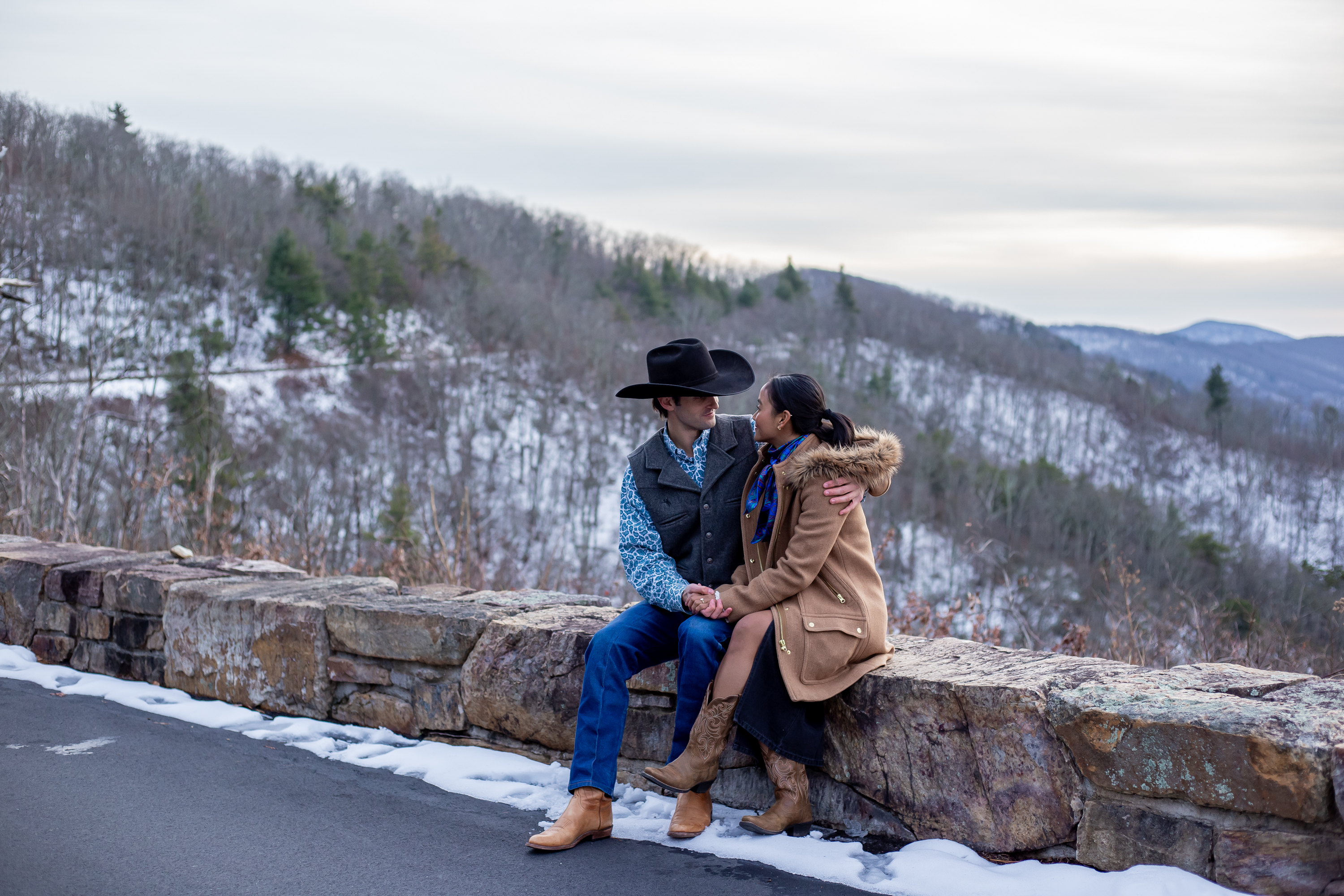 Couple in layered winter coats sitting together on a stone overlook wall at a snowy Skyline Drive overlook with snow-covered mountains behind them
